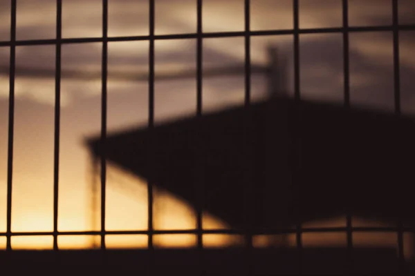 Silhouette of an industrial building behind a lattice fence against sunset yellow orange sky in the evening. Warehouse, factory building roof. Commercial property exterior. Wire fence. Industrial zone