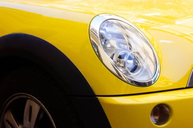 Oval front headlight of modern stylish yellow car. Sedan automobile MINI Cooper Countryman up close. Automotive business. Licensing, driving lessons, road travel concept. Bright cars details