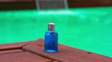 Cosmetic in plastic or glass blue mini bottle in travel version on wooden pier by swimming pool green water. Natural shower gel, shampoo, liquid soap in jar. Mockup facial skincare cosmetics product.
