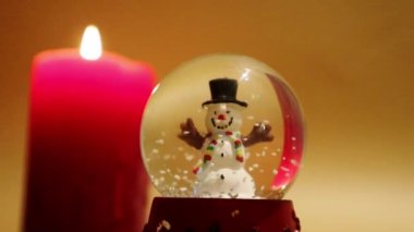 New Years glass toy, snow globe with cheerful snowman inside and snowflakes falling down. Home decor, red candlelight. Cozy Christmas atmosphere. Happy New year 2023 winter holidays wallpaper.
