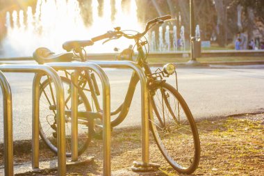 Classic two-wheeled bike parked in a bicycle parking lot in a city park on a sunny day at sunset in front of a fountain. An environmentally friendly mode of transportation for the urban environment.