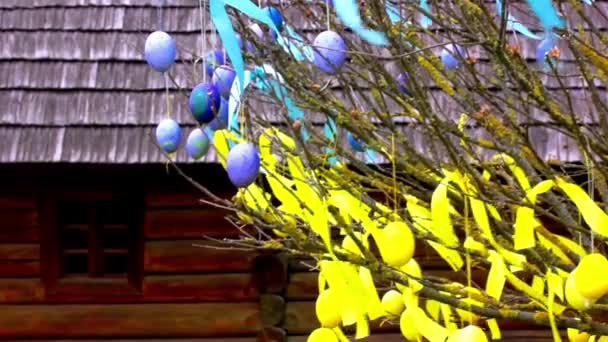 春の庭の果樹園の木の枝にぶら下がっている黄色と青のイースターエッグ 春の宗教休日のビデオ 木製のログハウスに対して屋外の通りにイースターの装飾 ウクライナの伝統 — ストック動画
