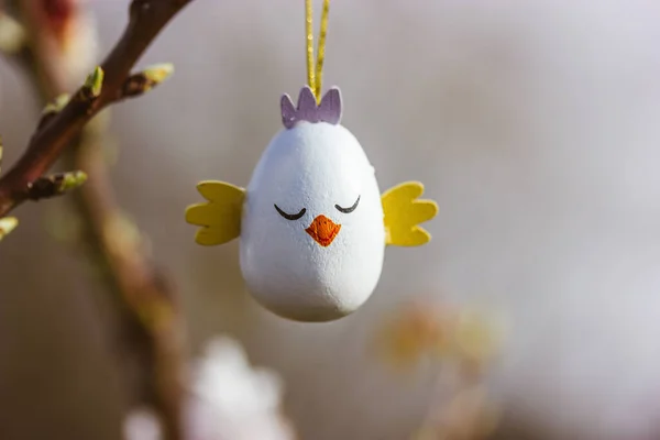 Easter egg in a form of a cute chicken hanging on a branch of a flowering tree in a spring garden in sunny day. Easter wallpaper for desktop. Greeting card. Decor for a springtime religious holiday.