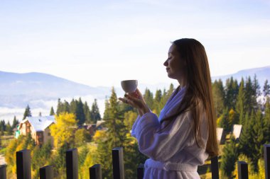 Genç, esmer, bornozlu bir kadın elinde çay fincanı, balkonda duran sıcak kahve içeceği. Kız temiz sabah havasını soluyarak eğleniyor. Tepeler, orman, dağlar. Spa eğlencesi, doğa rahatlaması.