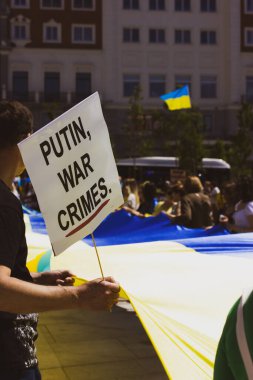 A white poster with slogan Putin war crimes in the hands of a protester at a rally in support of Ukrainians over the war unleashed by Russia. Stay with Ukraine. Ukrainian flag. Vertical photography. 