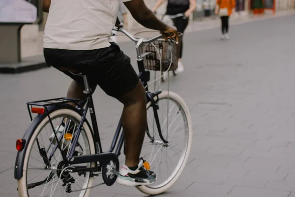A stylish modern dark-skinned hipster young man riding black white bicycle with basket on bike path in a summer city. Sporty lifestyle, leisure, sport, recreation in urban space. Free place for text.