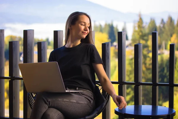Young woman, girl works at laptop sitting on terrace balcony in summer day. Remote online work home in a green forest. Happy entrepreneur, student girl smiling relaxing outdoors. Wireless technology.