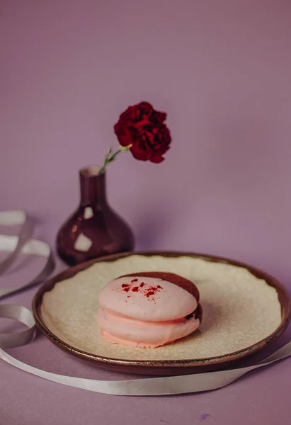 Pink brown chocolate whoopie pie on round plate on a table. Pink lilac carnation flower in vase still life. Sweet American cookie. Round trendy details in food photo. Soft biscuit cake for birthday.
