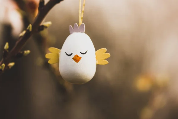 Easter egg in a form of a cute chicken hanging on a branch of a flowering tree in a spring garden in sunny day. Easter wallpaper for desktop. Greeting card. Decor for a springtime religious holiday.