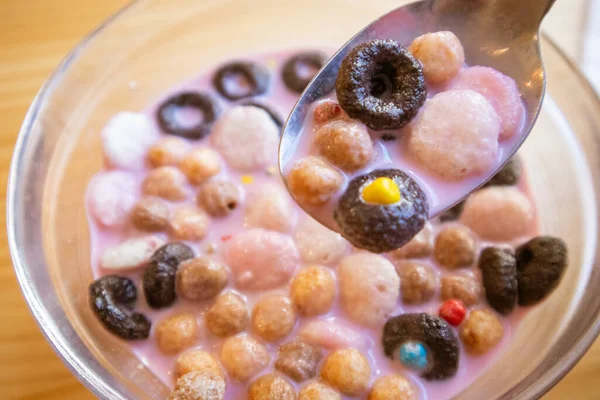Dry breakfast in glass round plate bowl on a wooden table in cafe. Delicious cornflakes drenched in pink strawberry milk. Healthy breakfast. Food for kids. Different type cereals, multicolored balls.