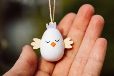 İnsan elinde dekoratif Paskalya yumurtası. Avucunun içinde beyaz tavuk şeklinde şirin, komik bir Paskalya oyuncağı kolye. Bahar mevsimi dini bayram kutlaması Nisan ayında. DIY el yapımı dekorasyon.