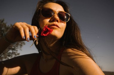 Yeşil bir tarlada elinde kırmızı haşhaş olan güzel bir esmer kadın. Bahar günü doğa yürüyüşüne çıkan bir kız. Kırsal turizm. Dünya günü. Dişi hippi yürüyüşçü gezgin hayat tarzını seviyor..