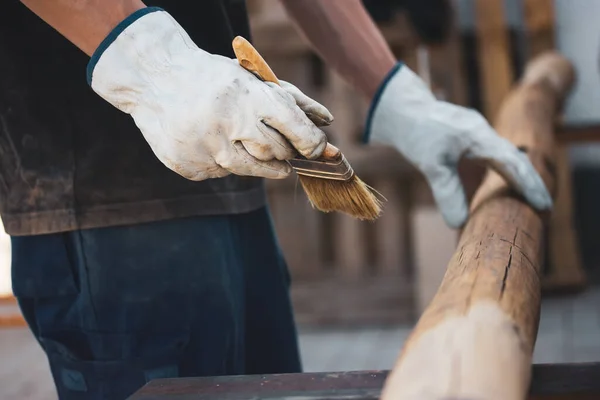 A faceless master carpenter works with wood by treating a surface with glue to protect it from insects. Carpentry work holding a brush. Products made of wood. Logs of trees. Wood staining, woodworking