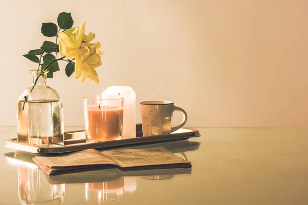 Still life in beige tones with a stylish tray, a cup of tea or coffee, a blooming yellow rosebud in a glass vase, an old open book lying on a table. Modern home decor for interior. Copy space for text