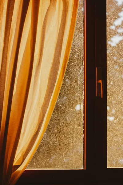 Odasında altın sarısı kahverengi bir perde olan ahşap bir pencere güneş ışığıyla dolmuş. Sıcak, sade bir atmosfer. Güneşli bir sabah ışığı. Kırsal kesimde bir evin ayrıntıları. Eski kirli pencere camı