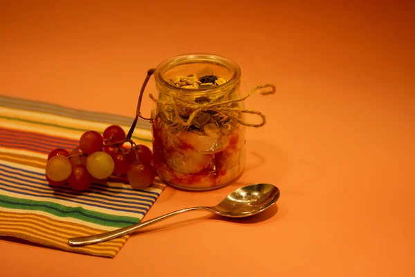 在桌子上的玻璃瓶里放着天然希腊酸奶一个金属甜点勺和一串葡萄 健康的早餐 带有橘红色背景的燕麦 天然食物 乳制品甜点 复制空间 — 图库照片
