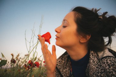 Şirin bir esmer kız kırmızı kokulu bir çiçeği kokluyor. Çayırda yetişen kır çiçeklerinin tadını çıkaran profilli bir genç kadının yüksek kaliteli fotoğrafı. Dişi kadın el çiçeğine dokunuyor.. 