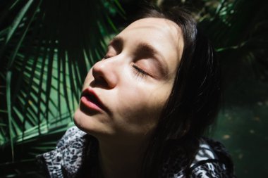 Palmiye yapraklarının arasında gözleri kapalı esmer bir kadının portresi. Güneşli bir günde palmiye ağacının altında bir kız. İnsan ve doğa birliği. Rüya gibi bir ruh hali. Tropikal bahçe ormanlarında turist Yüksek kaliteli fotoğraf