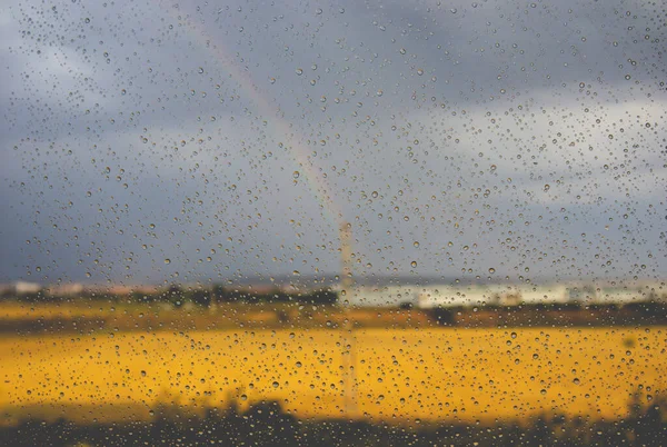 在阳光灿烂的雨天 窗外的彩虹 天气预报 窗玻璃表面的雨滴和景观 草地的景观 灰色的蓝天和黄色的草地 — 图库照片