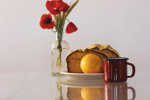 Rural still life with red poppies in glass vase and a plate with biscocho Bizcocho lemon muffin on kitchen table. Sweet dessert prepared at home. A red cup of coffee or tea. Biscotto pastries, cakes.