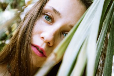 Bahçedeki yeşil tropik palmiye yaprakları arasında sakin genç bir kadın. Makyaj pembe rujlu bir kadın yüzü. Yorgun görünüyorsun. Güzel mavi gözlü bir kız. Kadın Psikolojisi. Korku ve Endişe