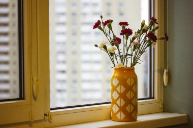 Küçük pembe ve beyaz karanfilli sarı vazo. Modern bir ev için çiçek dekoru. Pencere pervazında bir buket çiçek var. İçeride sıcak bir ışık altında. Bahar tatili için tebrikler. Çiçek hediyesi..