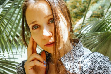 Güneşli bahar gününde yeşil tropik palmiye yaprakları arasında leopar desenli ceketli güzel bir kadın. Botanik bahçesinde gezen kadın turist. Gülümseyen sevimli bir yüz. İnsan ve doğa birliği kavramı. 