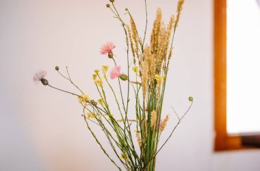 Vazodaki farklı kır çiçeklerinden bir buket beyaz duvara ve evin iç kısımlarındaki pencereye karşı. Ev dekorasyonu için çiçek aranjmanı. Kırsal evdeki kapalı alanlarda çiçek açan bitkiler. Bir odada çiçek açan yabani çiçekler..