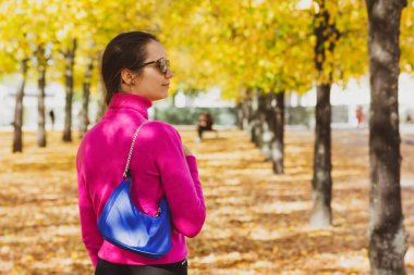 Berlin, İspanya. 2 Ekim 2022 Şık kadın, pembe kazaklı gülümseyen kız küçük mavi çantalı güneş gözlüğü takıyor, sonbahar parkında sarı yapraklara karşı poz veriyor..