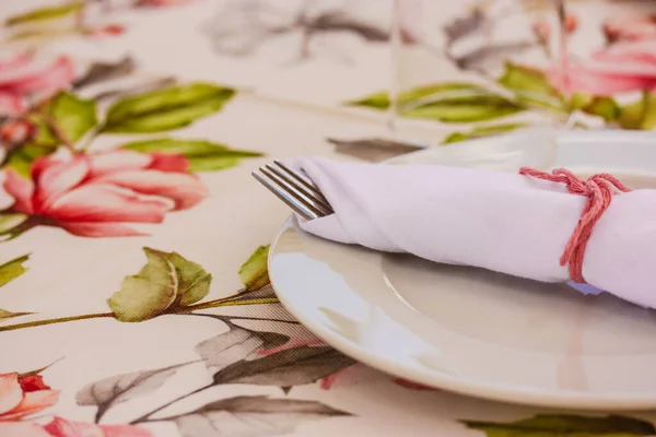 Elegantly table set at wedding, christening banquet. Floral tablecloth with pink flowers and white round plate, napkins. Thanksgiving holiday dinner decor. DIY napkin for birthday party lunch, brunch.