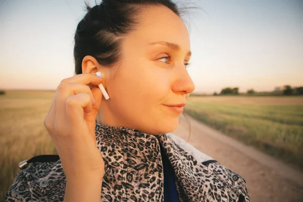 A young woman profile is listening a music and looking dreamily in distance walking in a countryside path meadow. Cute brunette girl on a walk with headphones in her ears. Female tourist in spring day