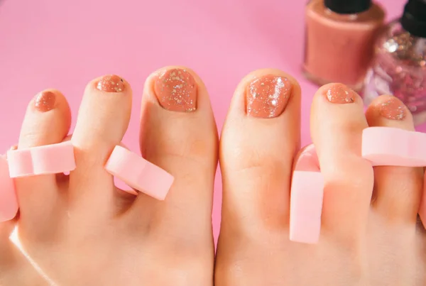 Women\'s feet with pedicure and pink toe separators on a background of nail polishes in the bottles. Beauty care for women concept. Pink women\'s pedicure and nails painted with glitter nail polish.
