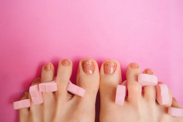 Women\'s feet with pedicure and pink toe separators on pink background. Beauty and care for women concept. Pink women\'s pedicure and nails painted with glitter nail polish.