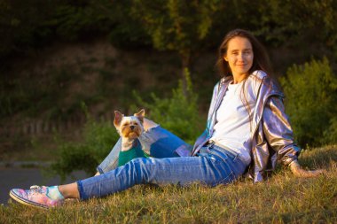 Baharın ilk baharında küçük bir köpek Yorkshire Teriyeri ile çimenlerde oturan mutlu güzel kız. Evcil hayvanları olan insanlar. Şık, genç, açık tenli, uzun saçlı, esmer bir kadın parkı rahatlatıyor..