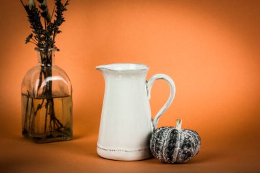 Beyaz bir su ve turuncu arka planda bir balkabağının yanında süt sürahisi ile sakin bir hayat. Cadılar Bayramı masa konsepti. Gıda hazırlamak için mutfak aletleriyle estetik kompozisyonu çiçekli cam vazo