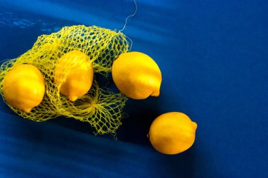 Sarı olgun limonlar mavi bir masa örtüsünün üzerine plastik bir torbadan düşerler. Modern moda iç mekan yiyecek arka planında güneşli yaz günlerinde sitruslu ağ alışveriş torbası Akdeniz yaz tatili