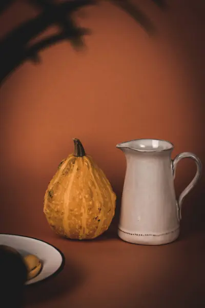 Rustic still life with a white water and milk jug next to a pumpkin on an orange background. Halloween table concept. Aesthetic composition with utensils for food preparation A glass vase with flowers