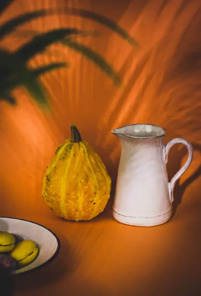 Rustic still life with a white water and milk jug next to a pumpkin on an orange background. Halloween table concept. Aesthetic composition with utensils for food preparation A glass vase with flowers
