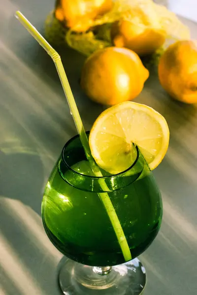 Green glass with refreshment water and yellow ripe lemons. Refreshing drink with vitamin C. Vertical food and drinks backdrop. A cocktail with citruses taste on a green table on a summer sunny day.