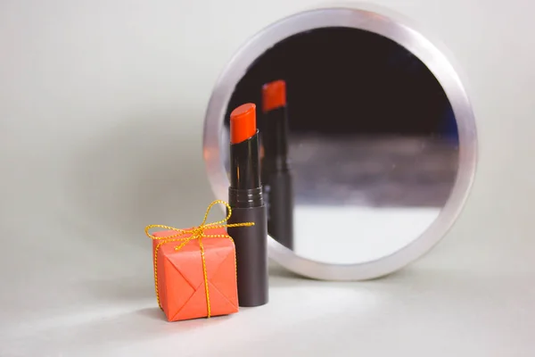 Women\'s makeup. Red lipstick and a round mirror with reflection on makeup table. Cosmetic still life on a contrast grey background. Decorative cosmetics sample and a red gift box, present box.