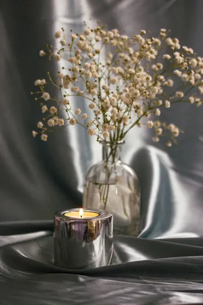 A burning candle in a metal candle holder on a table covered with a silver silk. Luxurious stylish decor for modern interior. A glass vase with small white summer flowers indoors. Floral postcard.