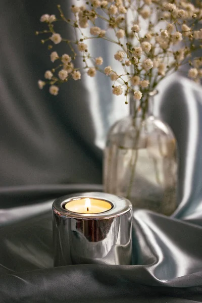 A burning candle in a metal candle holder on a table covered with a silver silk. Luxurious stylish decor for modern interior. A glass vase with small white summer flowers indoors. Floral postcard.