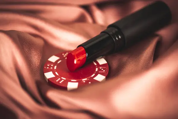 Red lipstick and casino chip on silk terracotta pink coral silk fabric. Female manipulation, seduction, flirting, romance, sexuality, fantasy, game, hunting on men. Lady cosmetic table. Love game.