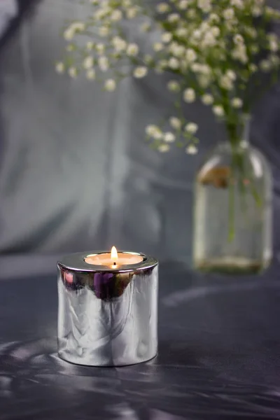 A burning candle in a metal candle holder on a table covered with a silver silk tablecloth. Luxurious stylish decor for modern interior. White flowers in glass vase at stylish home decor, decoration.