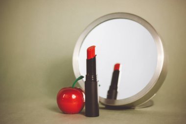 Makyaj masasındaki yuvarlak aynanın yanındaki siyah tüpte kırmızı ruj. Gri arka planda kozmetik ürünleri. Dekoratif kozmetik örneği. Kırmızı elmanın kokusu ve tadı olan rujun yansıması..