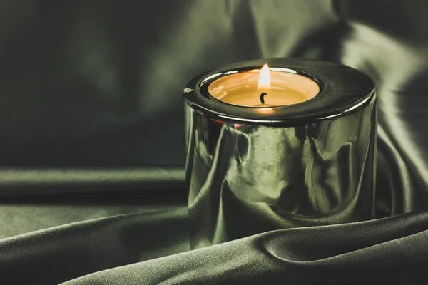 A burning candle in a metal candle holder on a table covered with a dark olive green silk tablecloth. Luxurious stylish decor for modern luxury interior. Decoration, spa, relax, meditation concept.