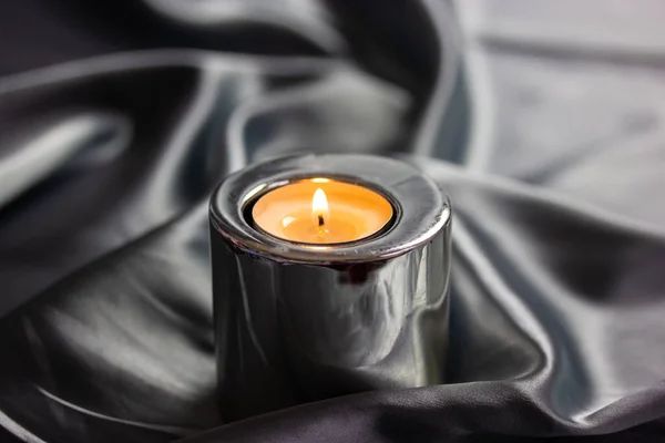 A burning candle in a metal candle holder on a table covered with a silver silk tablecloth. Luxurious stylish decor for modern interior A concept of tranquility, relaxation, meditation, self-knowledge