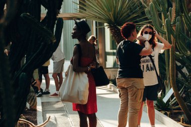 Madrid, İspanya. Ağustos, 12023. Botanik bahçesindeki insanlar, tropik çiçeklere, bitkilere, kaktüslere, sulandırıcılara bakan seralar. Koyu tenli, kırmızı elbiseli ve şapkalı kadın, doğayı keşfeden diğer insanlar..