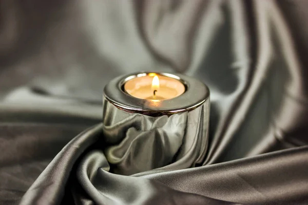 A burning candle in a metal candle holder on a table covered with a silver silk tablecloth. Luxurious stylish decor for modern interior A concept of tranquility, relaxation, meditation, self-knowledge