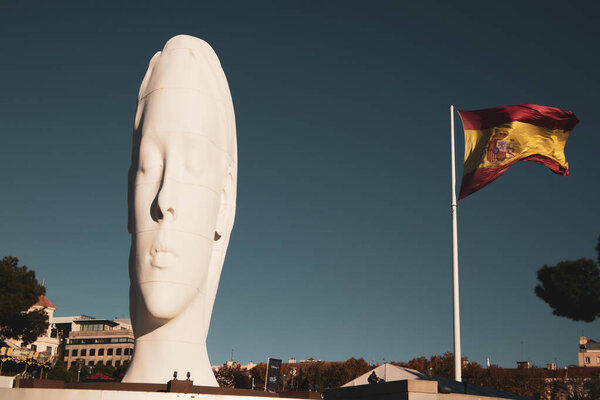 Madrid, Spain. November 22 2023 High huge white sculpture of young woman with eyes closed against blue sky. Masterpiece by artist Jaume Plensa on Colon square. Spanish flag on a wind. Dreamy girl face